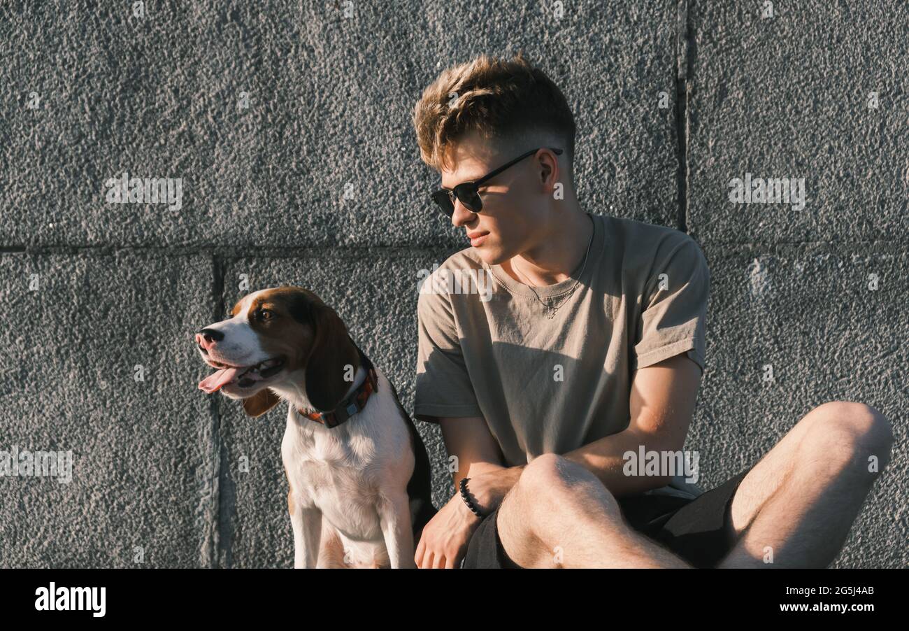 Young man and beagle dog sit together, concrete urban background. Lifestyle with pets, cute puppy and his owner Stock Photo