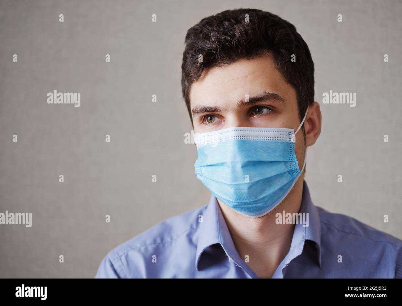 Sad man in a medical mask on his face in a coronavirus pandemic looks to the side - Concept of the new norm - A guy in a mask on his face on a gray ba Stock Photo