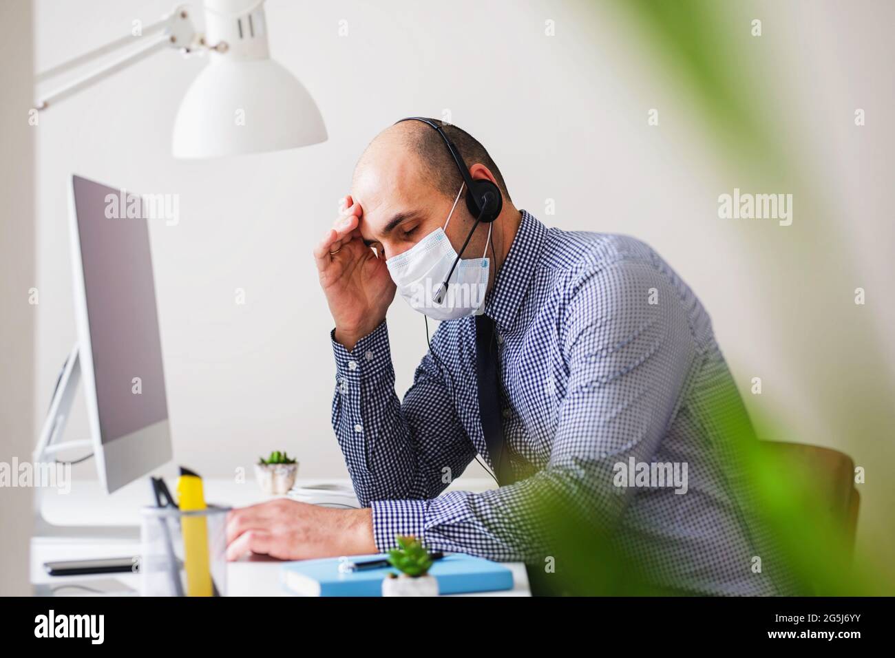 A man in a mask and with a headset suffers from headaches and fatigue - Depression at a call center employee under conditions of coronavirus - Stress Stock Photo