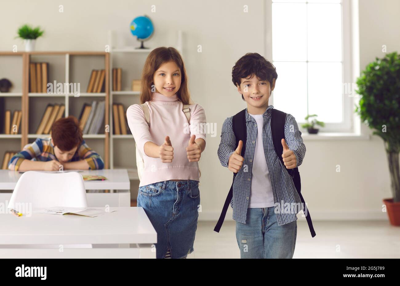 Boy and girl elementary school students show thumbs up standing in school classroom. Stock Photo
