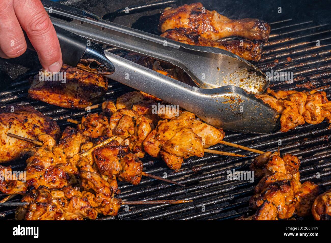 Cooking tongs turning Chicken pieces on skewers on a barbeque. Stock Photo