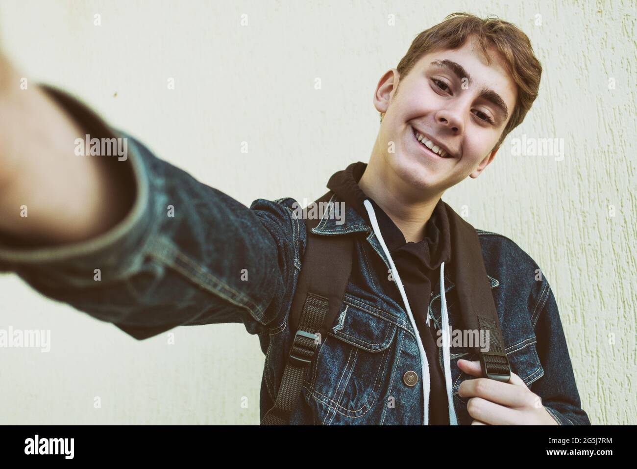 Boy teenager takes a selfie on the background of a light yellow wall - Happy teenager takes pictures of himself on the phone camera and smiles - Stude Stock Photo