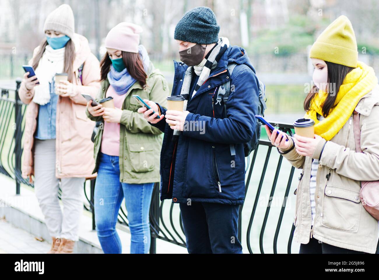 A group of young people stand at a distance from each other and keep their distance during the coronavirus epidemic - Millennials are typing on smartp Stock Photo