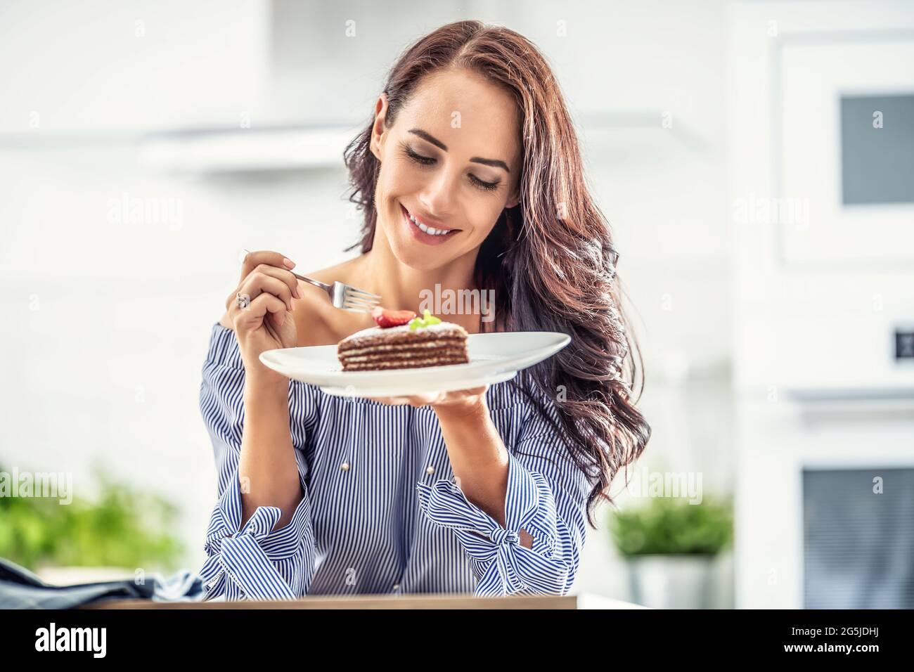 Layered brown adn white cake with strawberry and mint leaf on top held by a woman with a fork indoors. Stock Photo