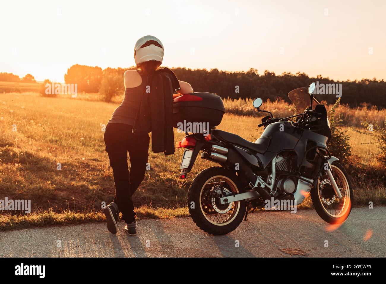 Female biker standing next to her bike, taking a break. Looking towards the sun. Afternoon light, copy space Stock Photo