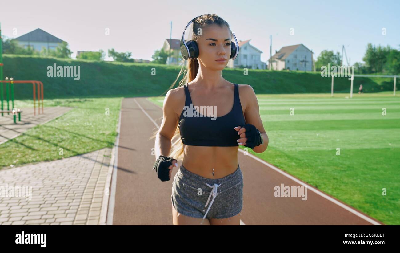 Front view of young athlete woman wearing big wireless headphones and black gloves running outdoors. Stunning girl with pony tail practicing jogging at stadium in sunny summer morning. Stock Photo