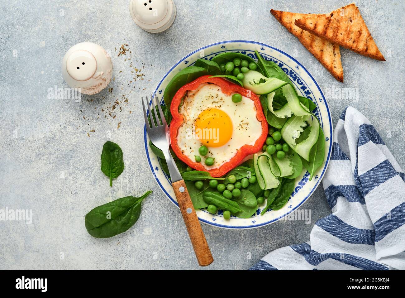 Red bell peppers stuffed with eggs, spinach leaves, green peas and microgreens on a breakfast plate on light grey table background. Top view. Stock Photo