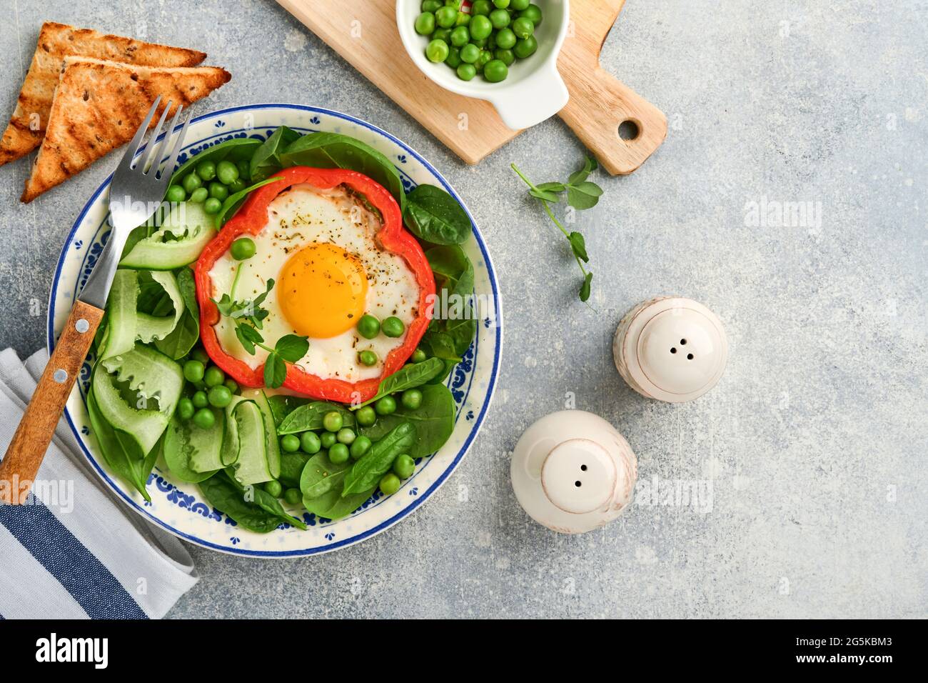 Red bell peppers stuffed with eggs, spinach leaves, green peas and microgreens on a breakfast plate on light grey table background. Top view. Stock Photo