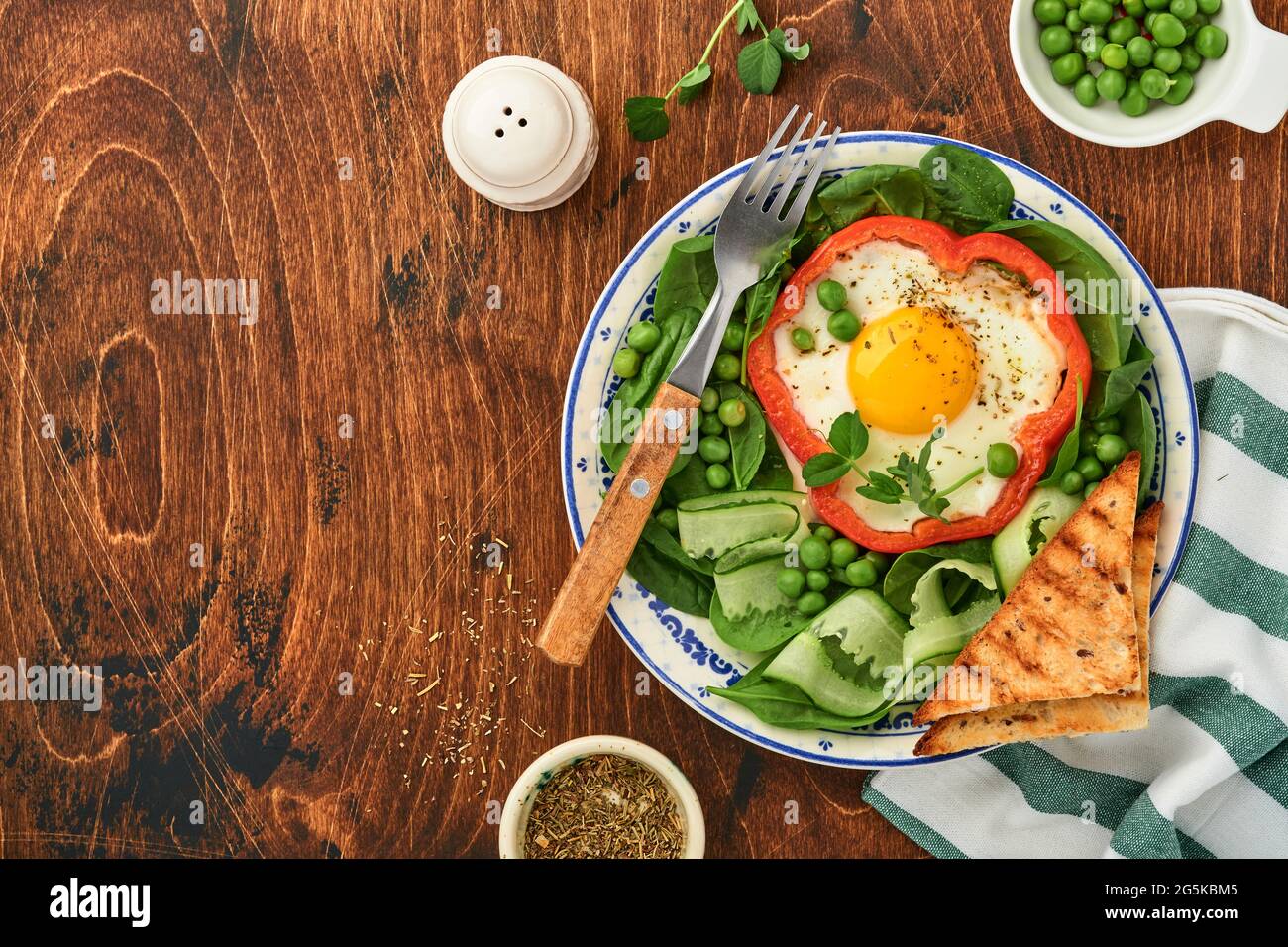 Red bell peppers stuffed with eggs, spinach leaves, green peas and microgreens on a breakfast plate on old wooden table background. Top view. Stock Photo