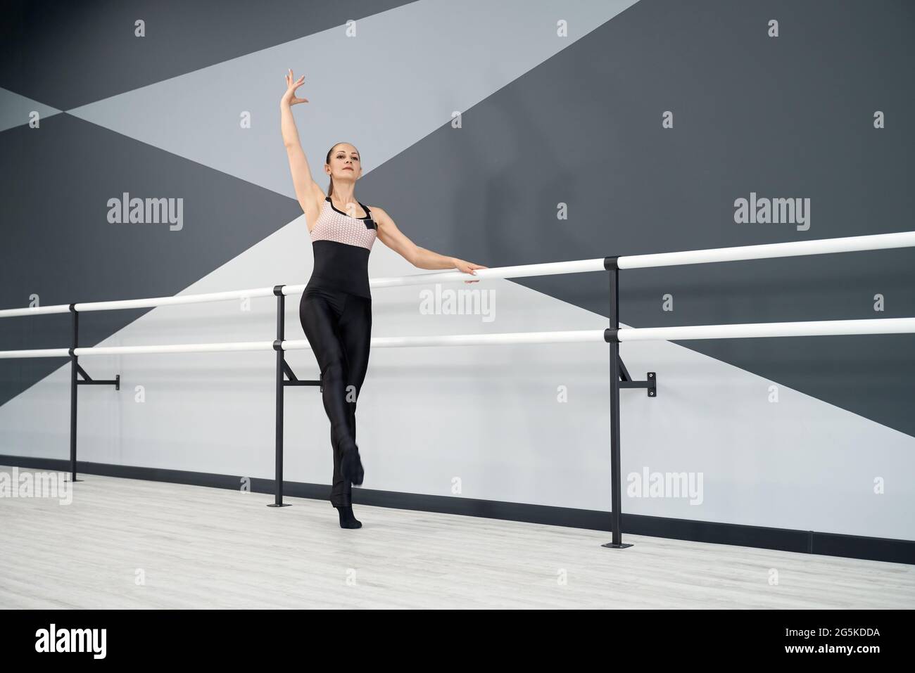 Front view of sportswoman in tight outfit posing with hand up, holding handrails in ballet studio. Graceful female adult athlete training in empty hall. Concept of choreography, gymnastics. Stock Photo