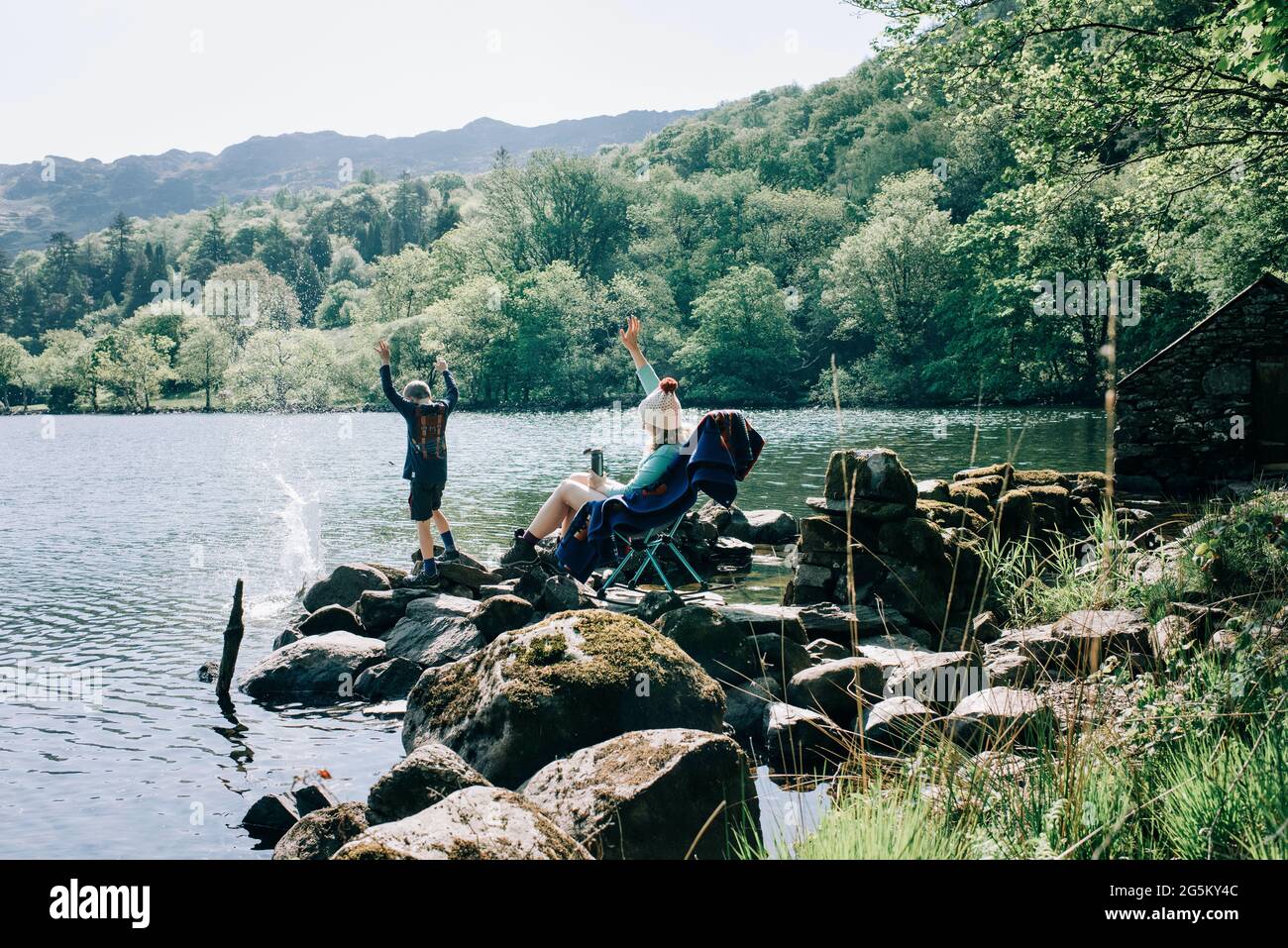 woman sitting on rocking chair by a lake whilst her kids throw rocks Stock Photo