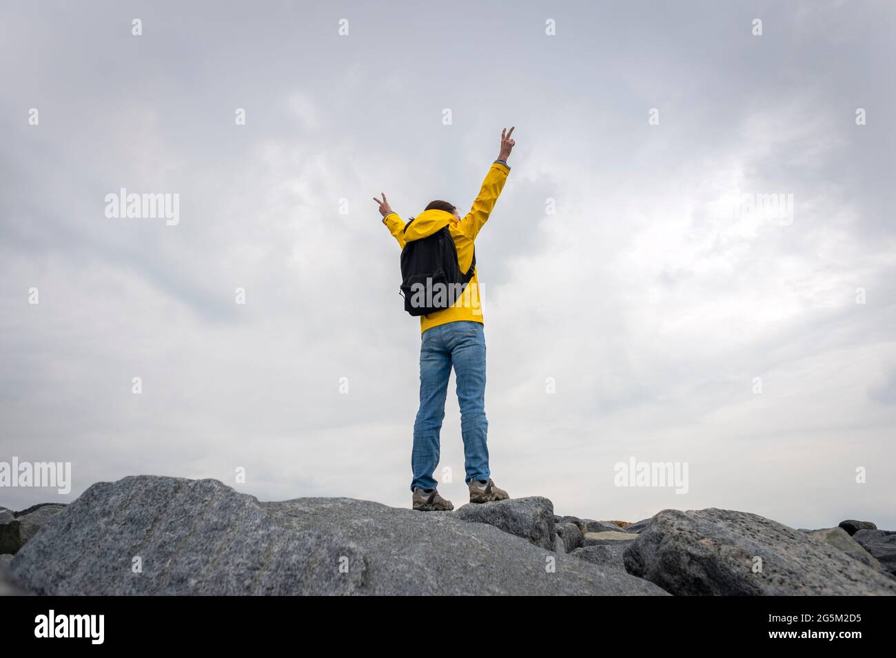 Back view of a person wearing a yellow outdoor jacket on the summit of rocks with arms raised in achievement. Stock Photo