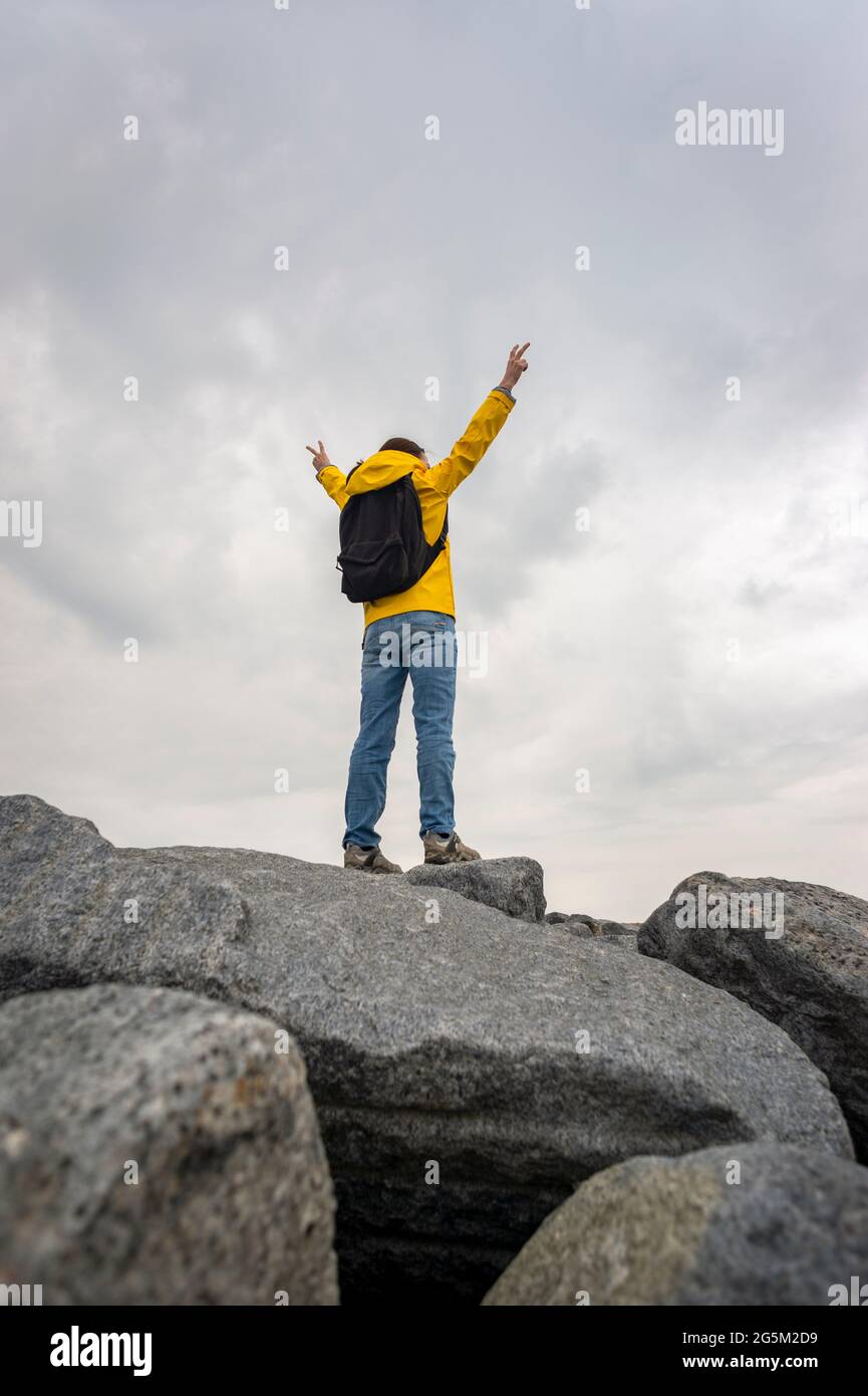 Back view of a person wearing a yellow outdoor jacket on the summit of rocks with arms raised in achievement. Stock Photo