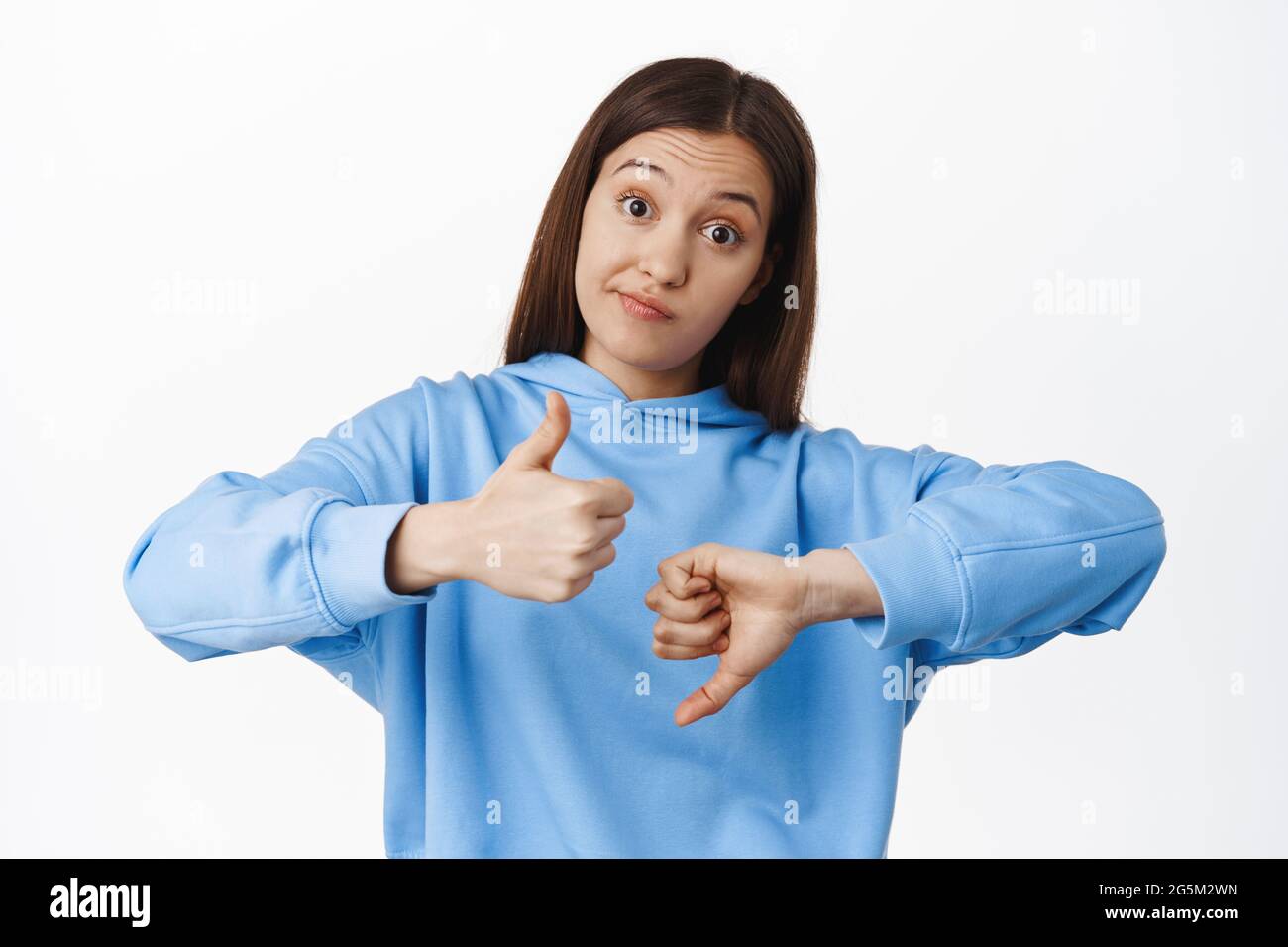 Not bad. Young woman smirk, shows like dislike, thumbs up and thumbs down gesture, medium rating, average result, standing in hoodie against white Stock Photo