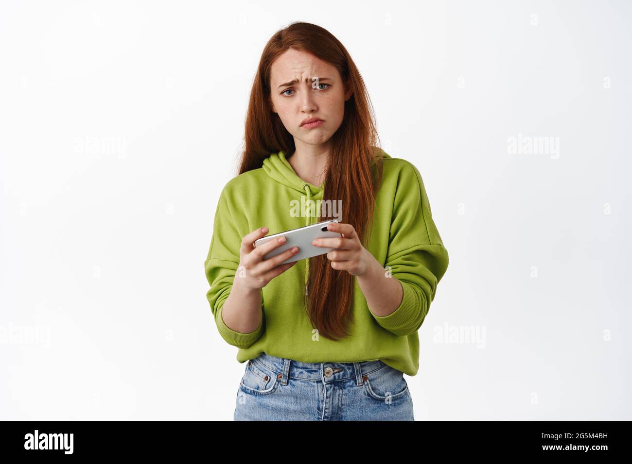 Sad redhead girl losing video game on mobile phone, watching upsetting video on smartphone and grimacing, frowning disappointed, white background Stock Photo