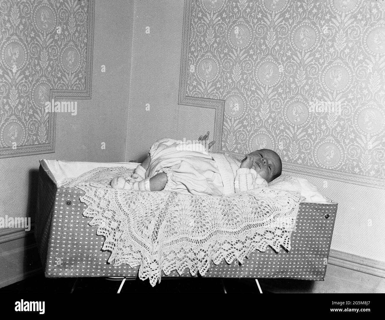 1960, historical, in a corner of a room, a baby resting on a small decorative knitted throw on top of a light travel cot of the era, England, UK. Stock Photo
