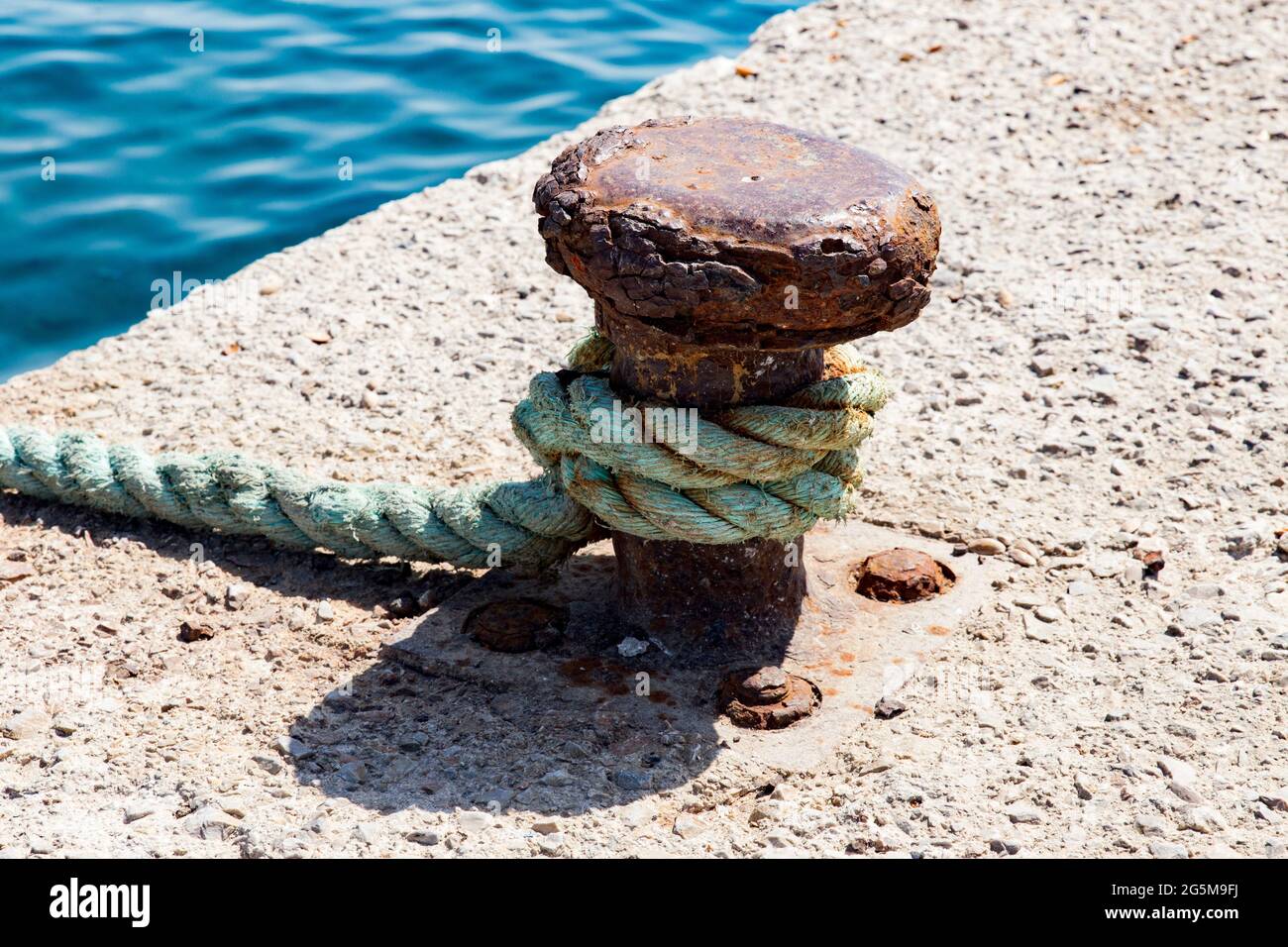 Rusty bollard with a strong rope attached under strong mediterranean sun with sea, high angle view Stock Photo