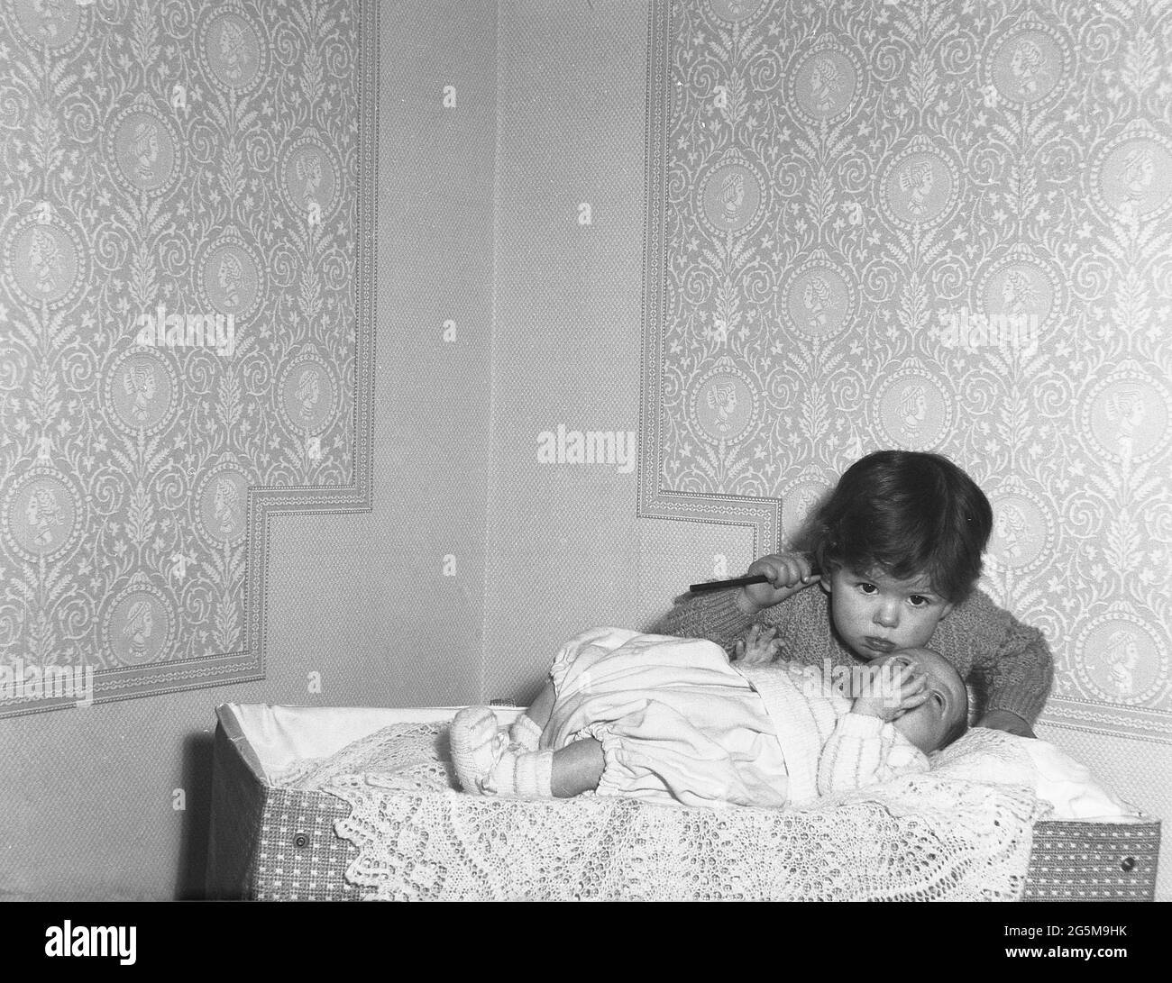 1960s, historical, in the corner of a room, a little girl checking on her baby sister lying on a throw on top of a travel cot of the era, England, UK. Stock Photo
