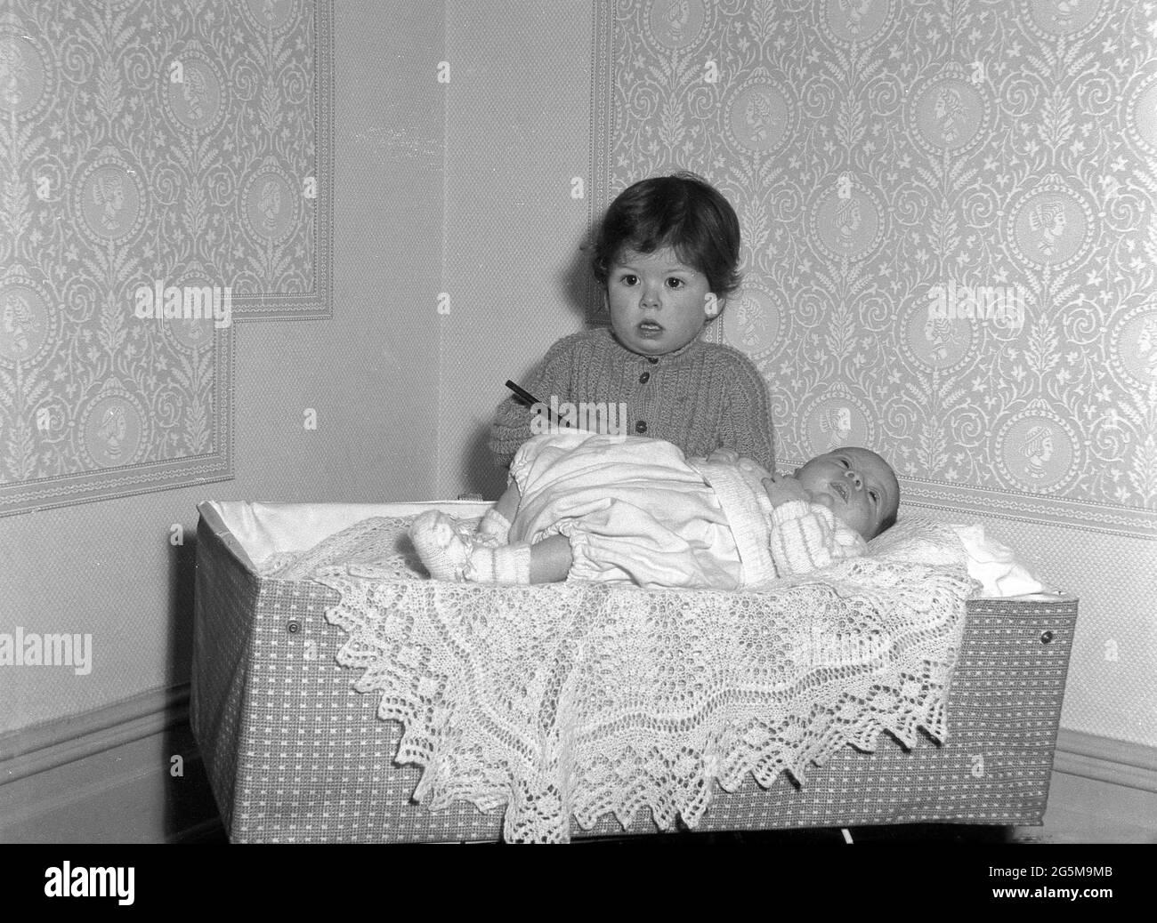 1960s, historical, in a corner of a room, a little girl standing beside a baby resting on a lace throw on top of a travel cot of the era, England, UK. Stock Photo