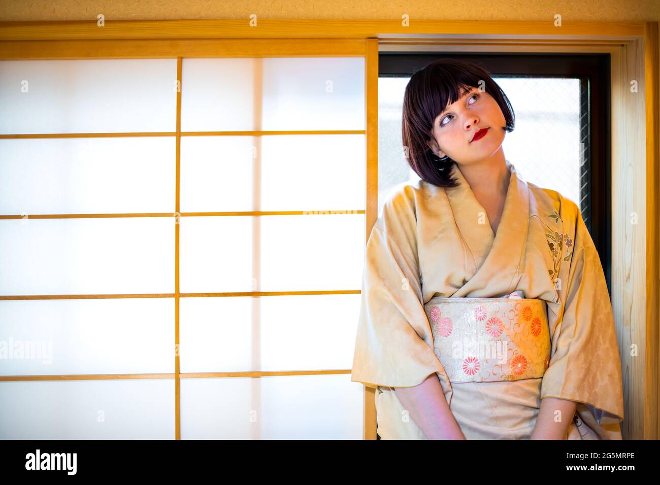 Traditional Japanese house with woman in kimono costume dress looking up with short black hair on windowsill of wooden sliding paper window Stock Photo