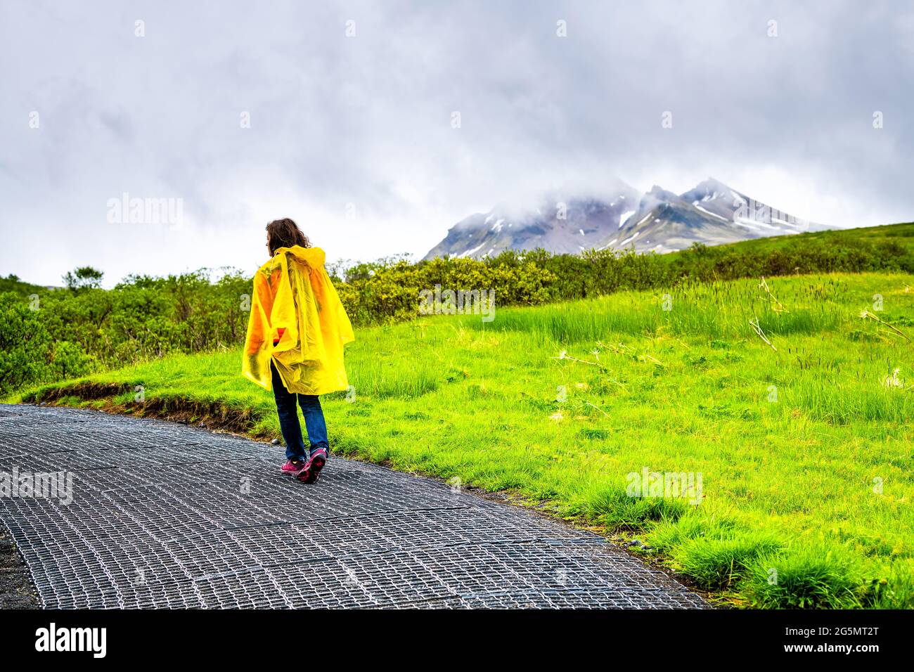 Skaftafell, Iceland green summer lush landscape and woman tourist in yellow poncho walking hiking on wet trail path hiking road rainy day in mountains Stock Photo