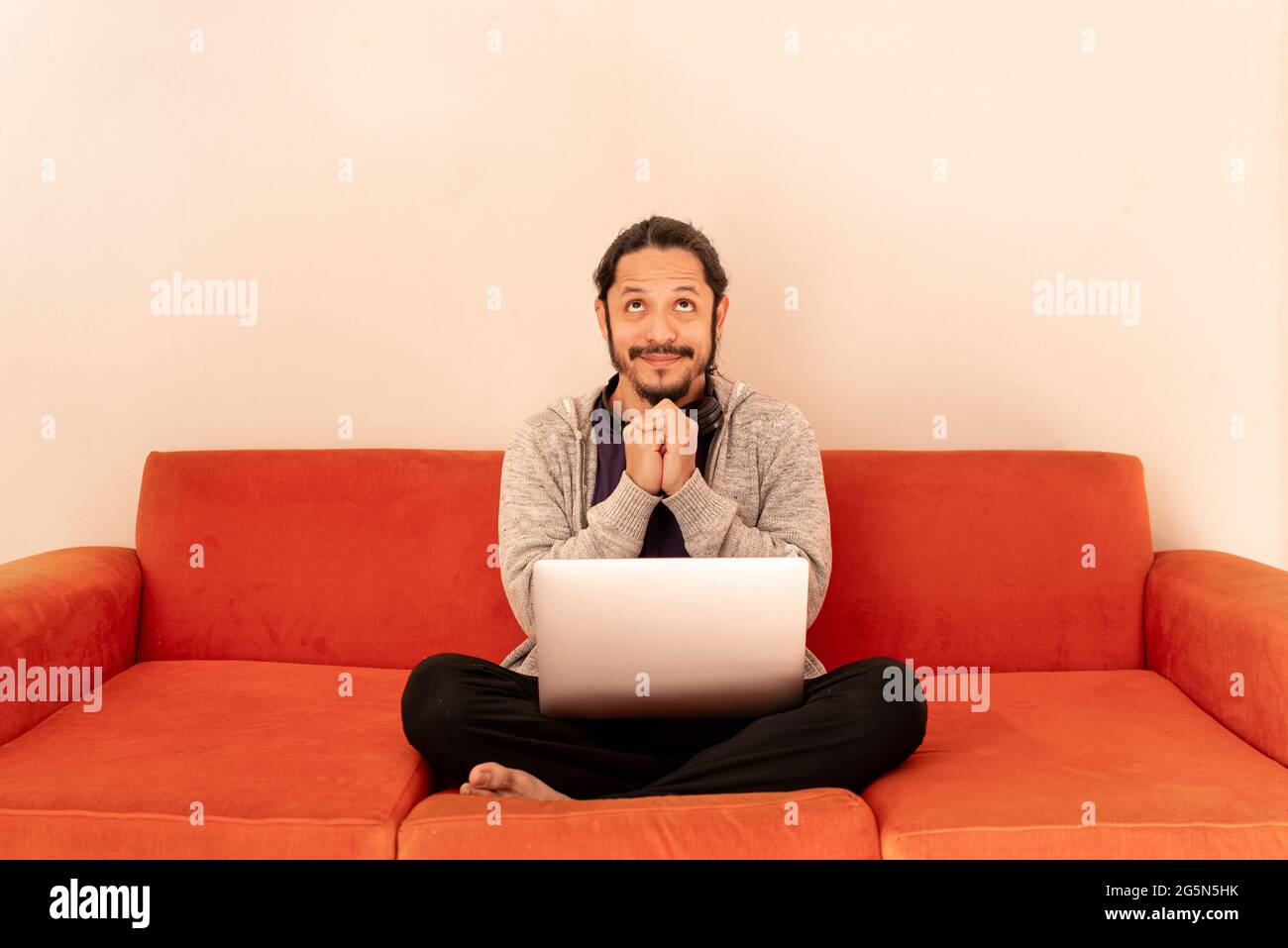 Young man looking up with funny face sitting on an orange couch sofa in the living room at home. Concept for sales time. Stock Photo