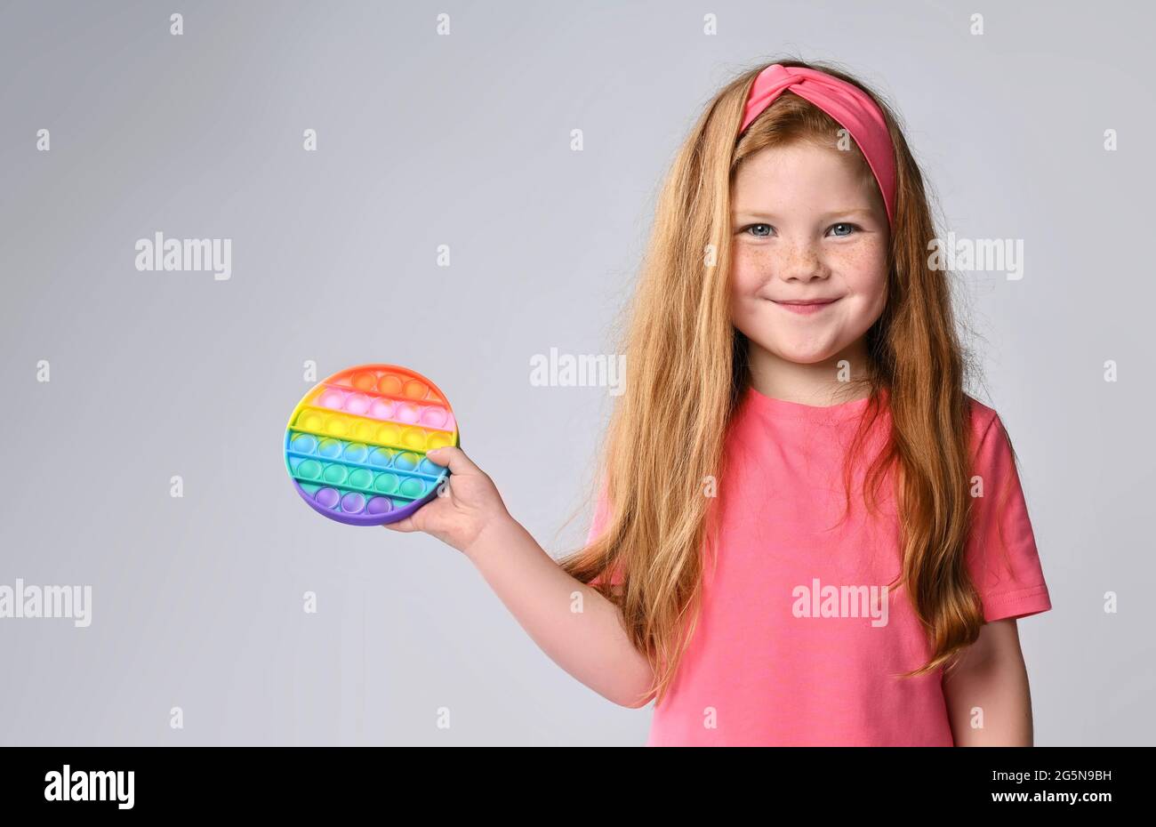 Cute, smiling kid girl in pink t-shirt demonstrates, shows her new trendy sensory toy, rainbow color round - pop it Stock Photo