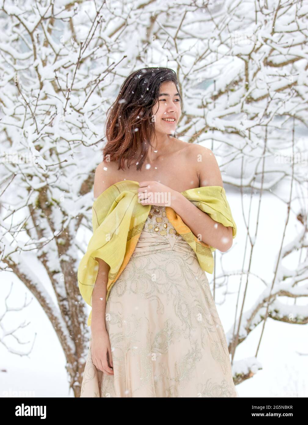 Biracial teen girl in formal gold colored dress and shawl outside on snowy day with snow covered tree branches in background Stock Photo