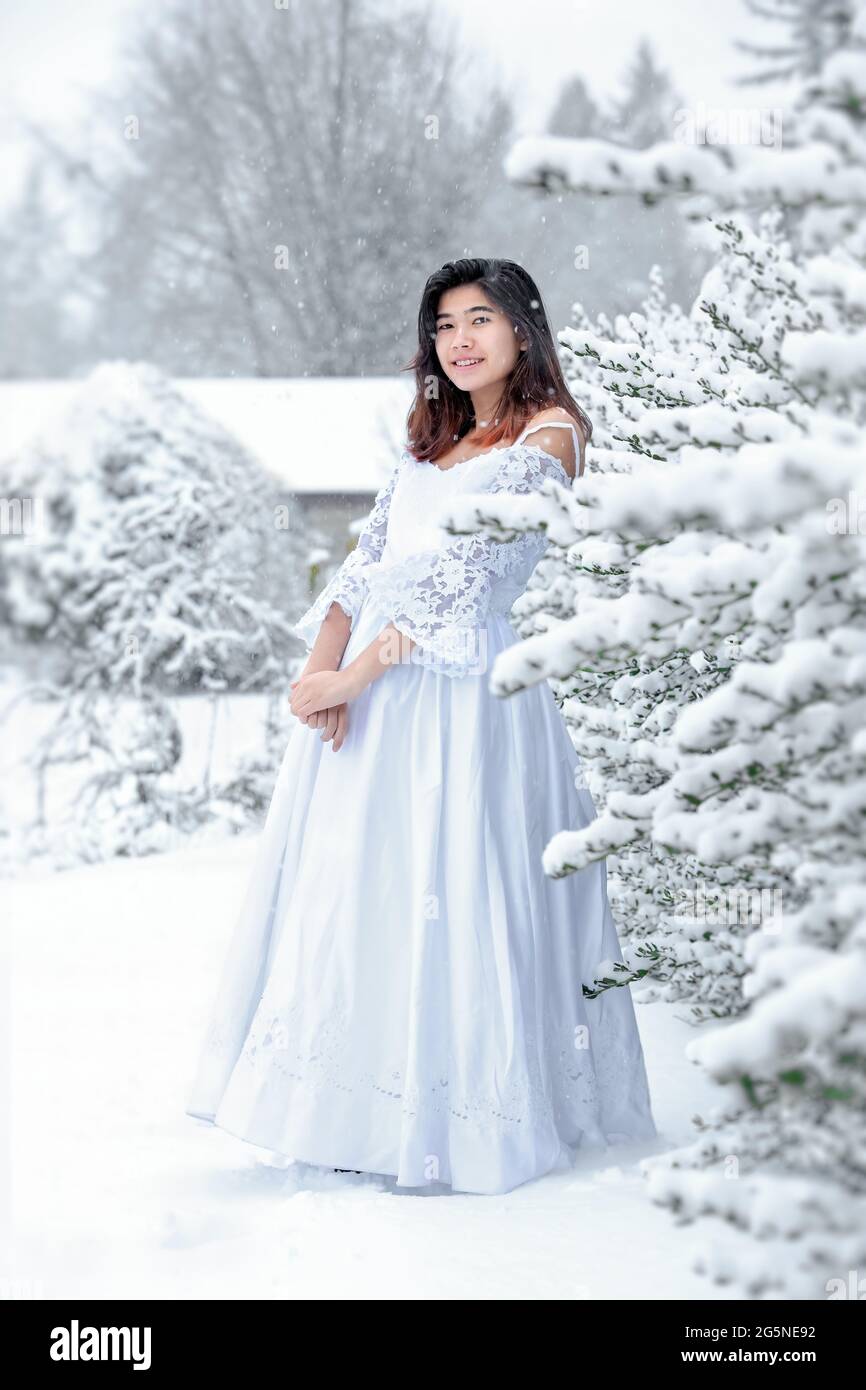 Biracial teen girl wearing lace white dress standing outside in snow during winter Stock Photo