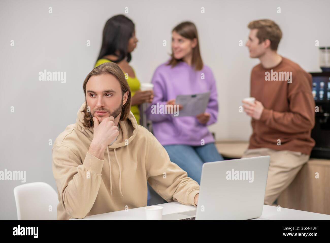 Thinking guy near laptop and colleagues chatting behind Stock Photo