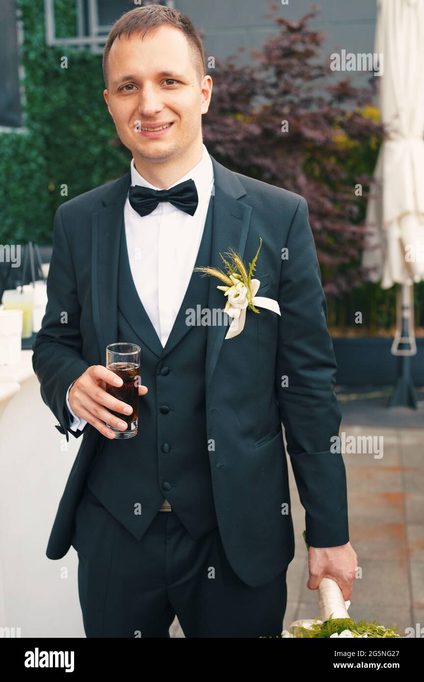 Handsome groom in formal suit having drink and holding bride's bouquet Stock Photo