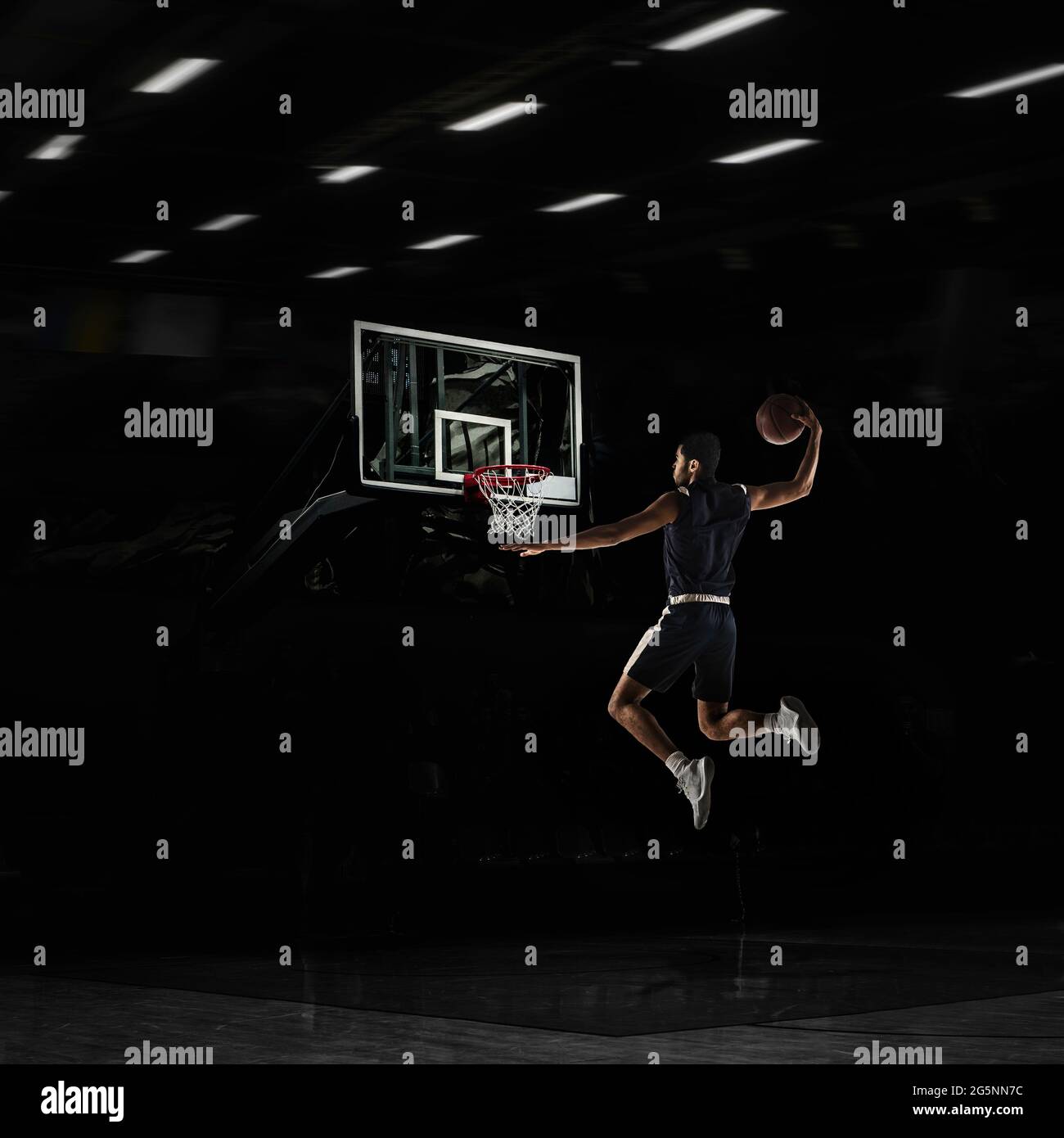 Young African sportsman, basketball player training in gym, idoors isolated on dark background. Concept of sport, game, competition. Slam dunk. Stock Photo