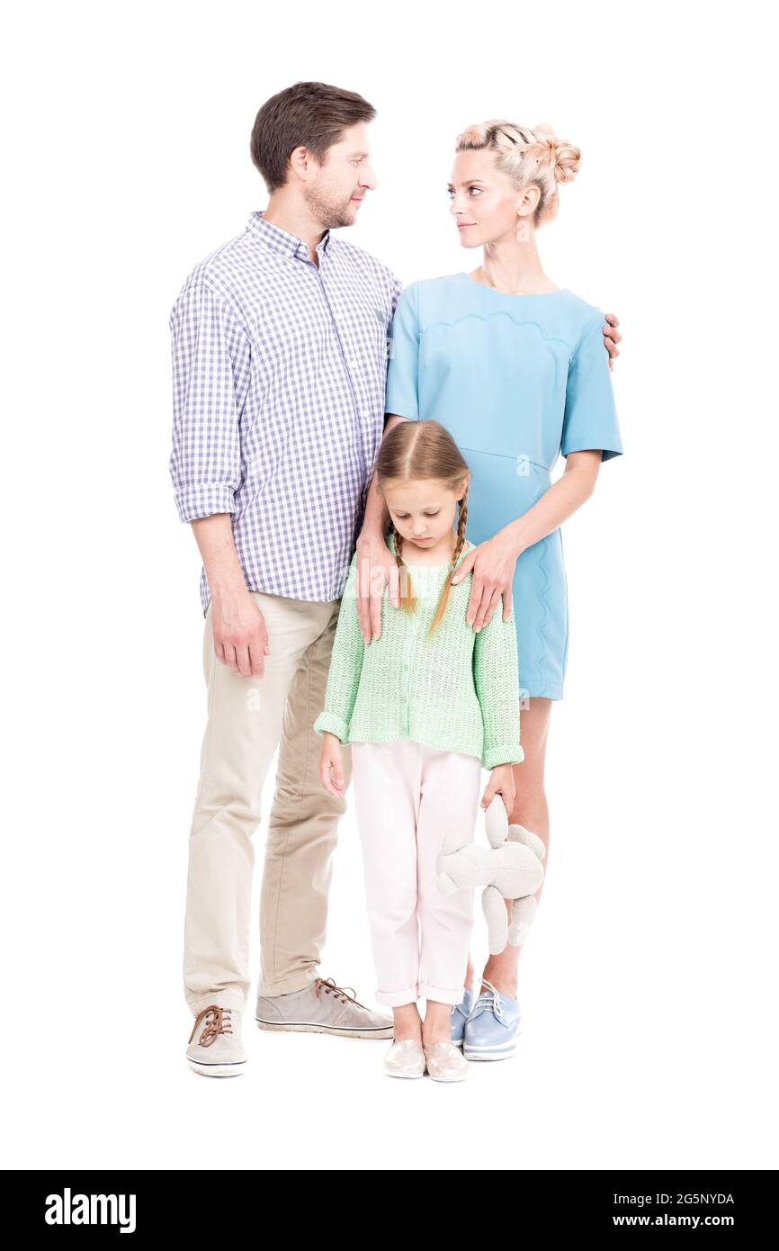 Vertical full length shot of young adult parents wearing casual clothes standing with their sad little daughter, white background Stock Photo