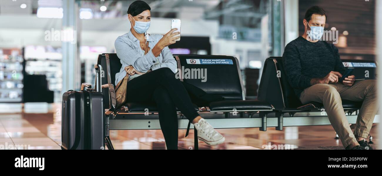 Man and woman traveler sitting apart maintaining social distance at airport. Male and female tourist with facemasks waiting at airport and using cell Stock Photo