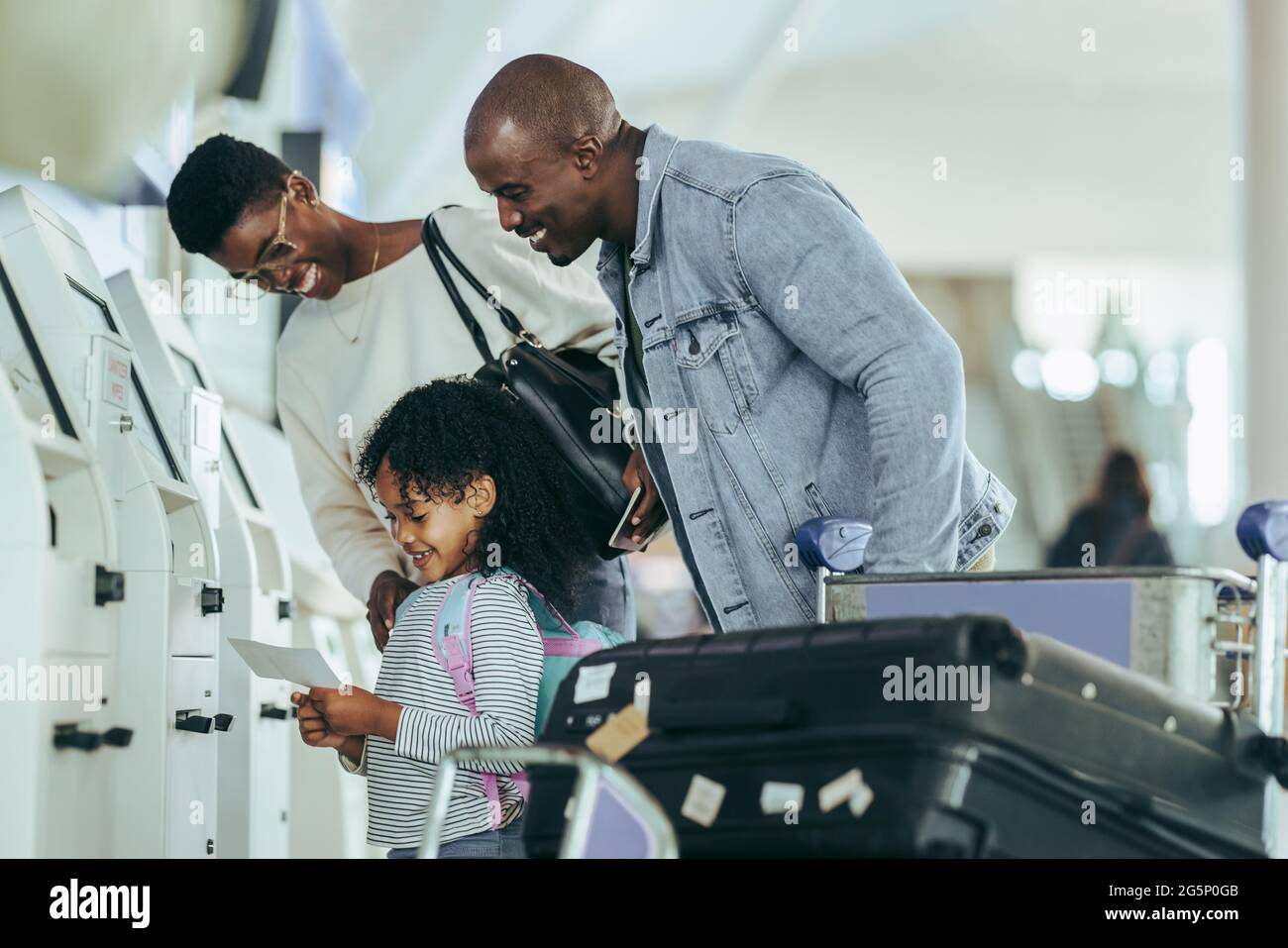 African family looking at the boarding pass in hands of their daughter at airport. Young girl looking happy to hold air ticket print while standing wi Stock Photo