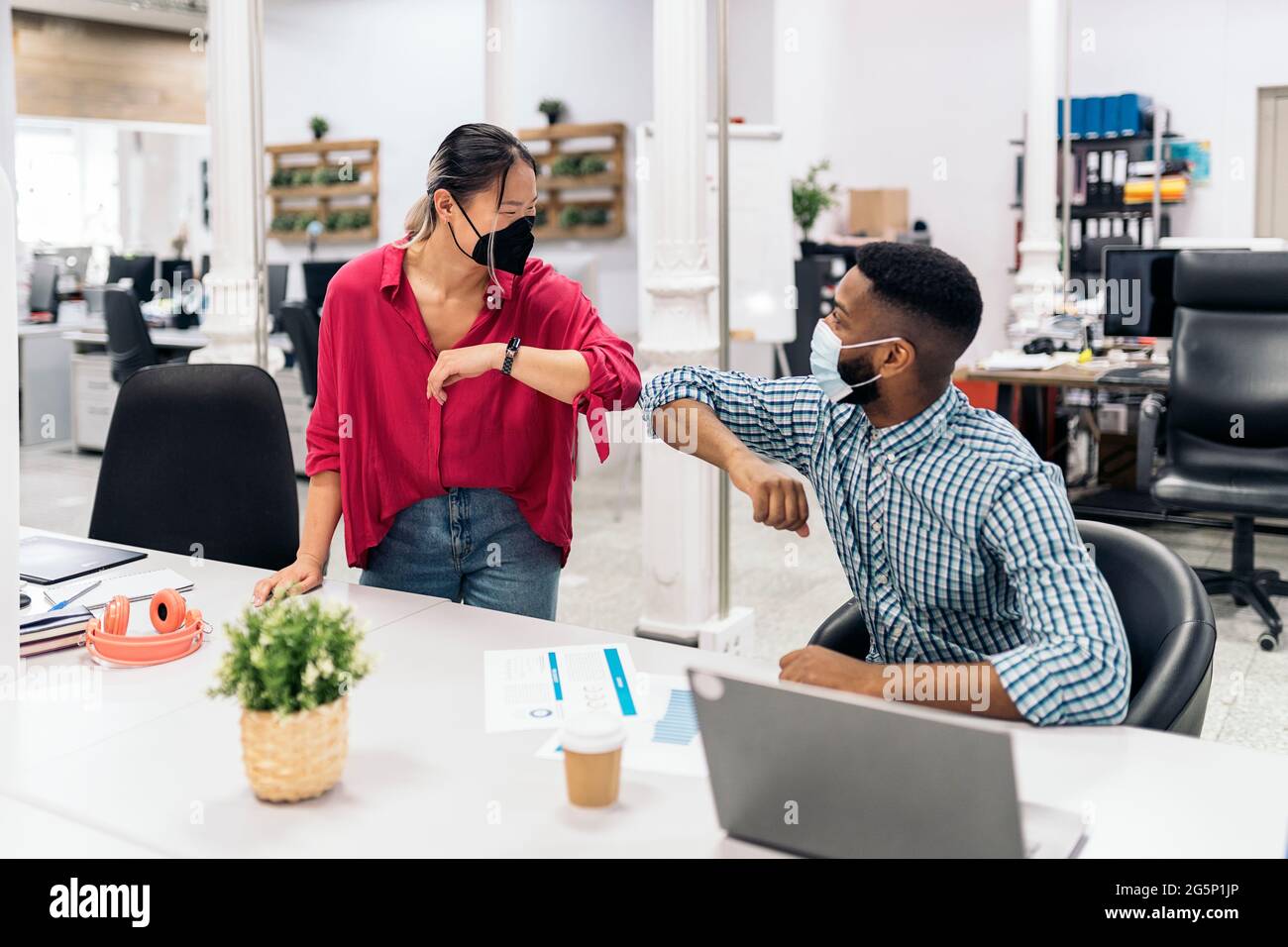 Young asian office worker talking with her african colleague and doing elbow bump. They are wearing face masks due to covid19. Stock Photo