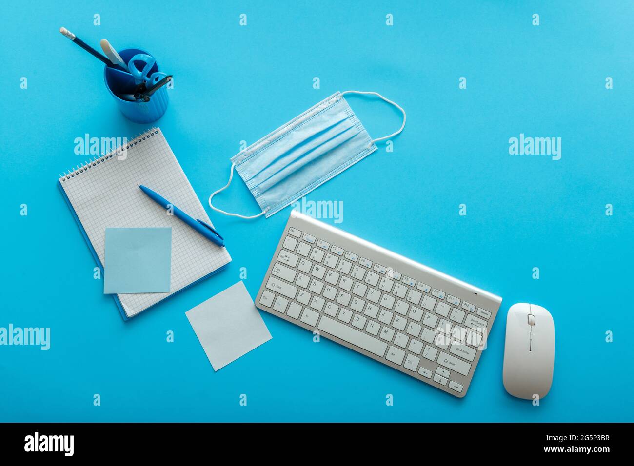 Back to school layout concept. Blue work desk with covid medical face mask, writes notes, notebook keyboard mouse at workplace. Desktop office space Stock Photo
