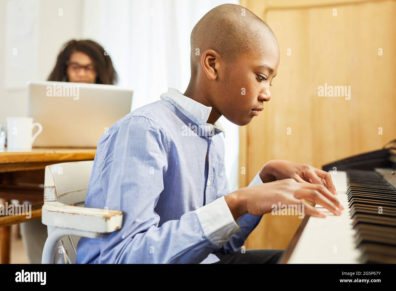 African boy learning to play piano in living room with mother on laptop pc  in background Stock Photo - Alamy