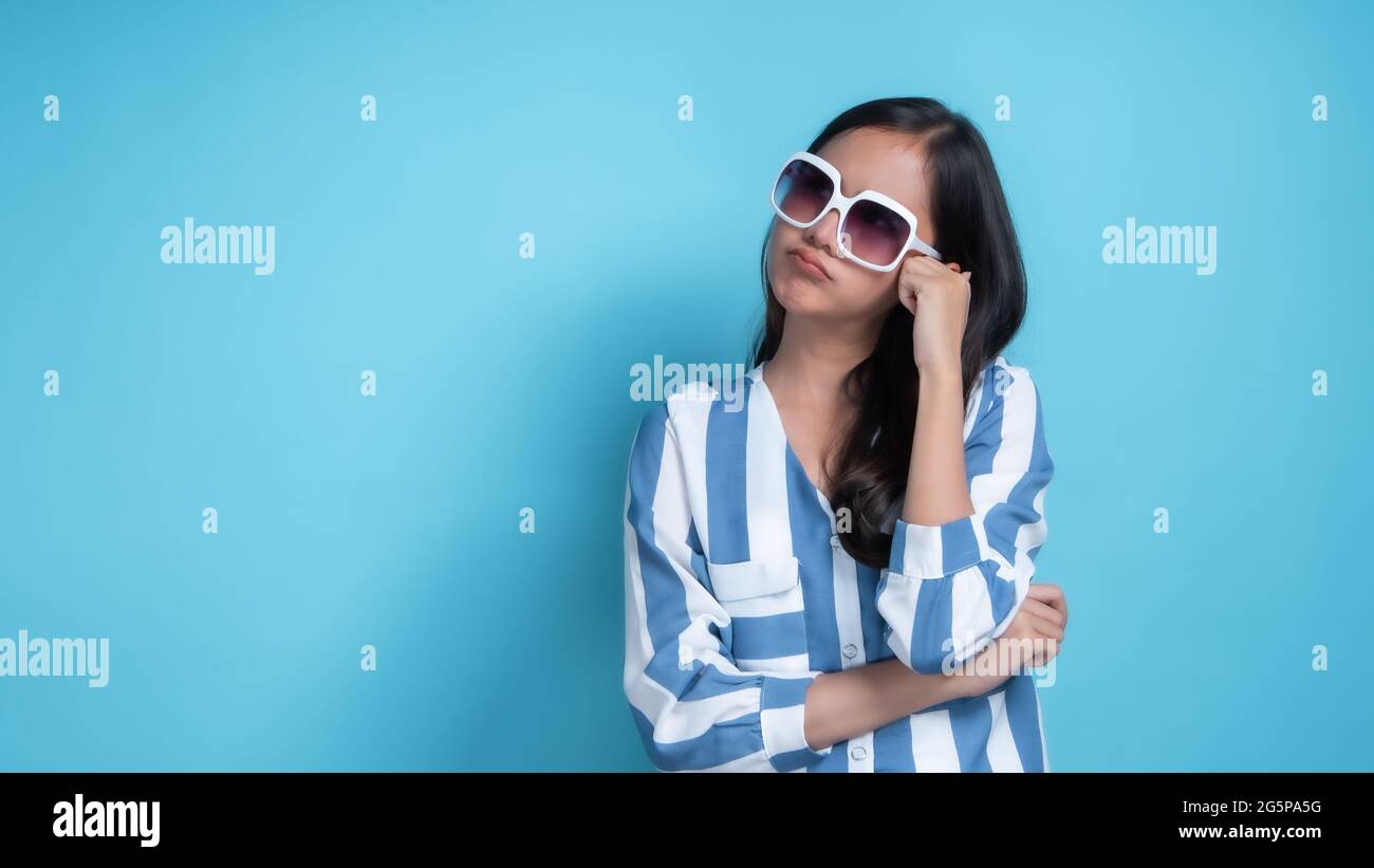 Portrait of young beautiful Asian woman wearing white glasses relax and innocent.cute girl think and consider somethings in studio on blue background. Stock Photo