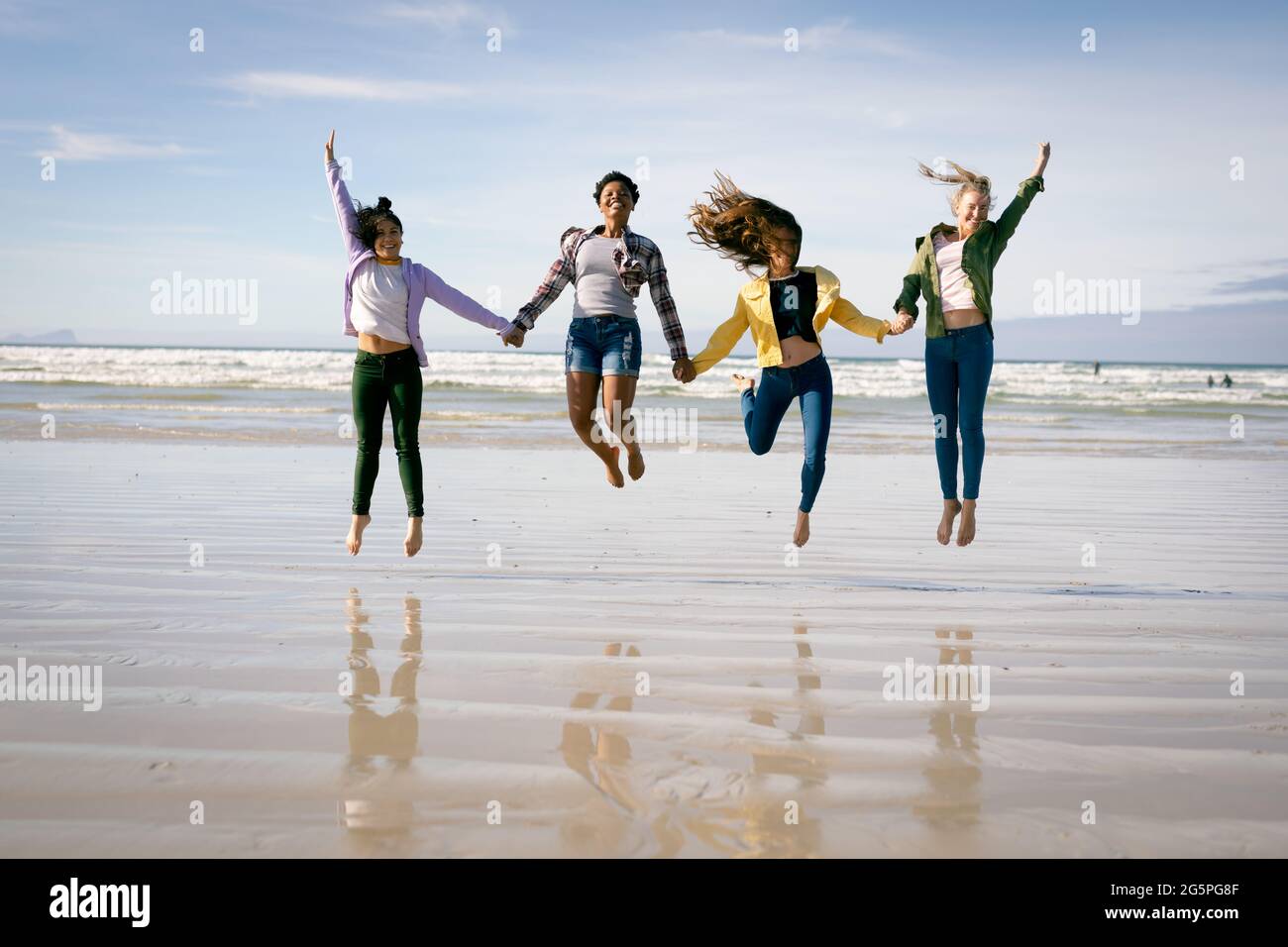 Happy group of diverse female friends having fun, walking along beach holding hands and jumping Stock Photo