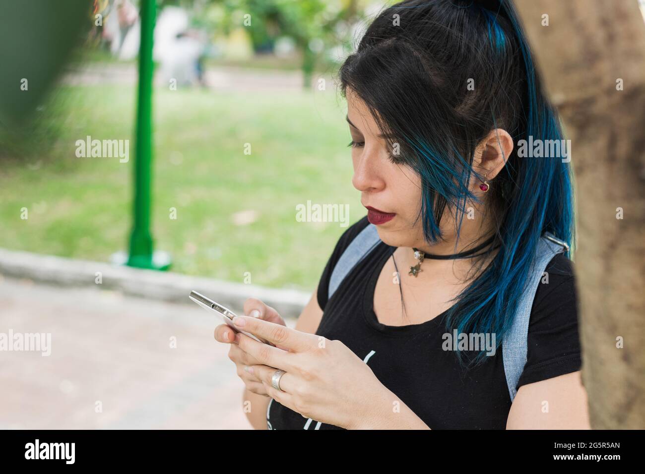 Beautiful young blue haired college girl looking at her social networks in a park leaning on a tree. girl looking at her cell phone in a park. surroun Stock Photo