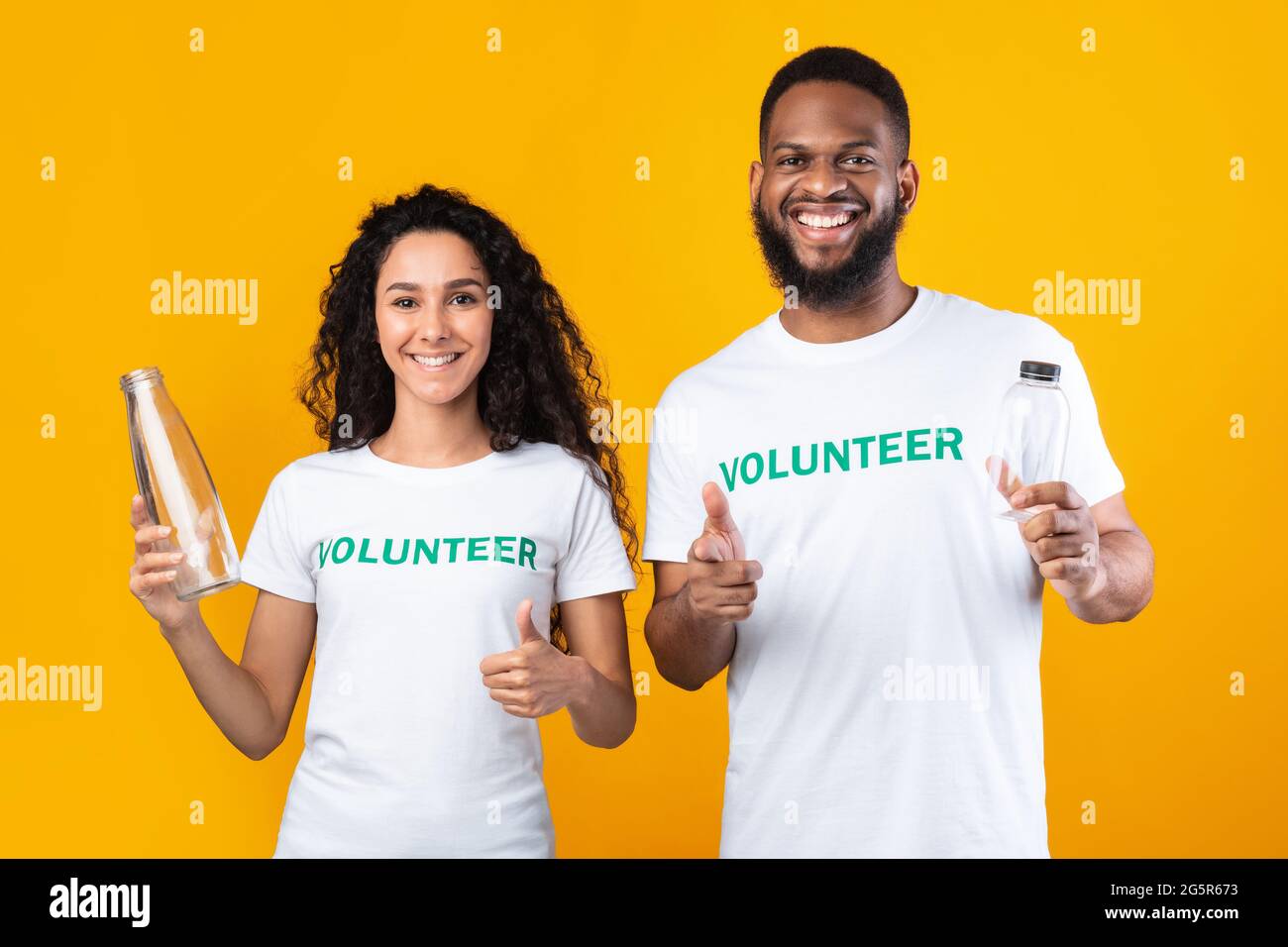 Two Volunteers Holding Glass And Plastic Bottles, Yellow Background Stock Photo