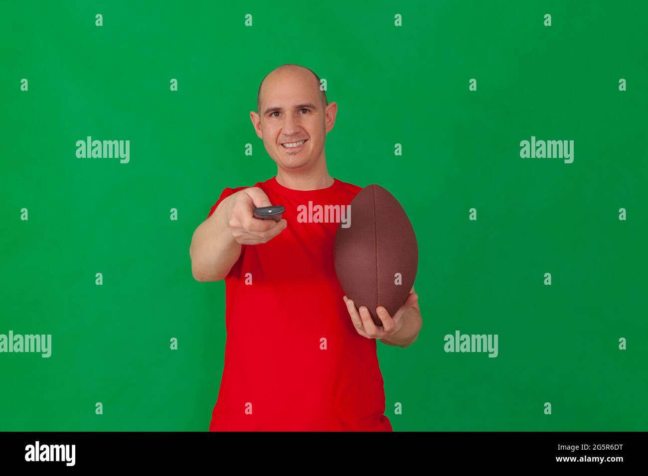 A bald caucasian man wearing a red t-shirt holds a soccer in his left hand and points a television remote control with his other hand. The background Stock Photo