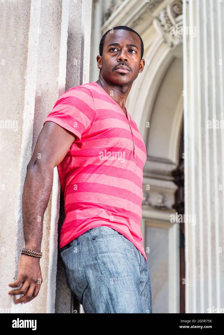 Young Man. Dressing in red, pink lines T-shirt,  gray pants, wearing a bracelet, a young black man is standing by a pillar, confidently looking forwar Stock Photo