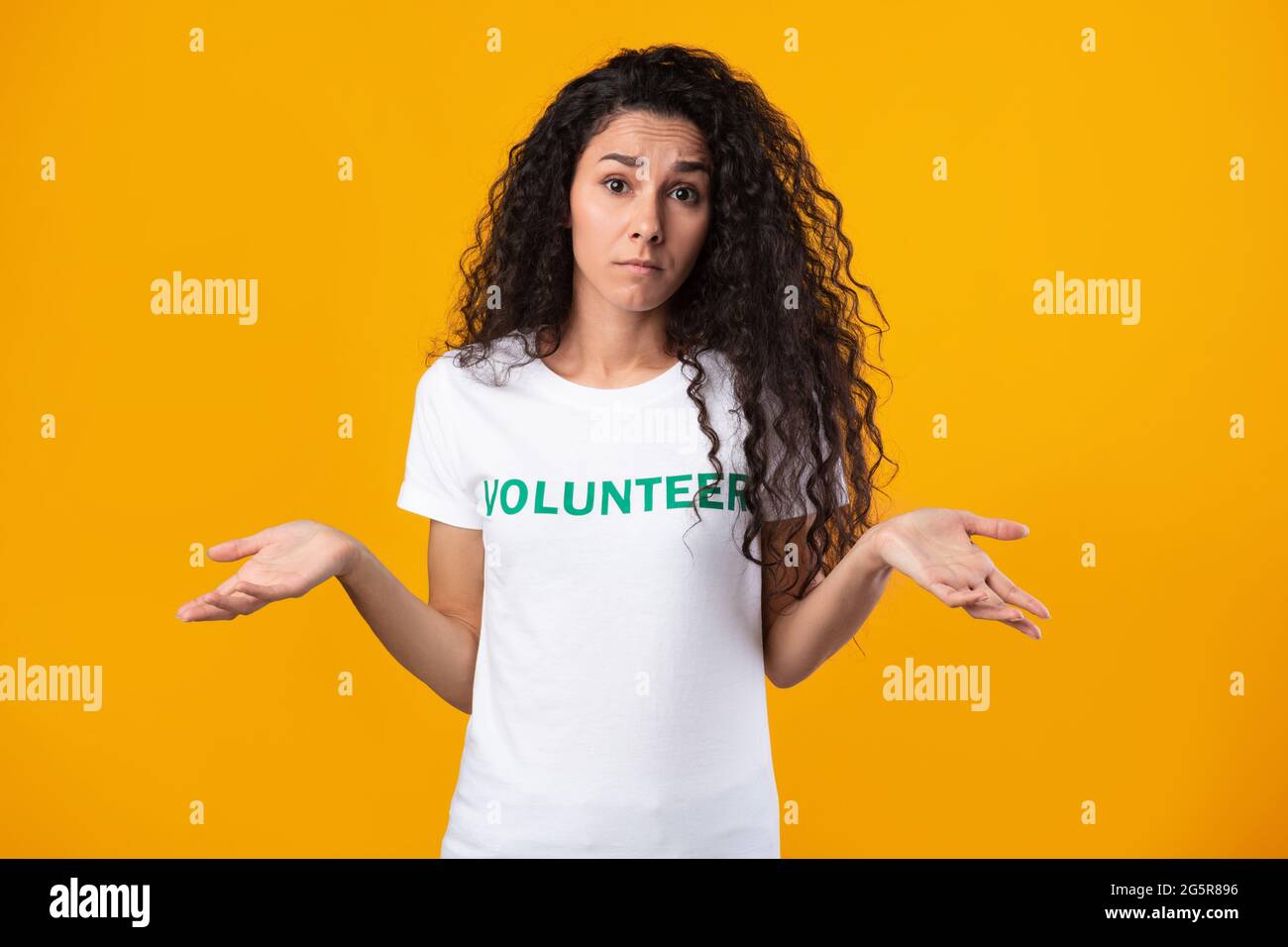 Puzzled Volunteer Lady Shrugging Shoulders Posing On Yellow Studio Background Stock Photo