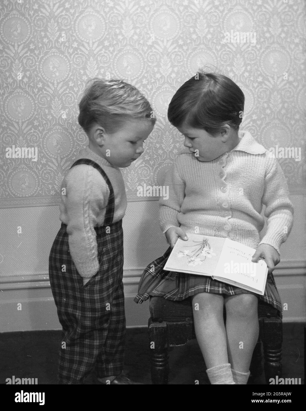1960s, historical, what's that book you're reading? a little boy in tartan dungrees showing interest in his sister's book she has on her lap, a picture book of flowers, England, UK. Stock Photo