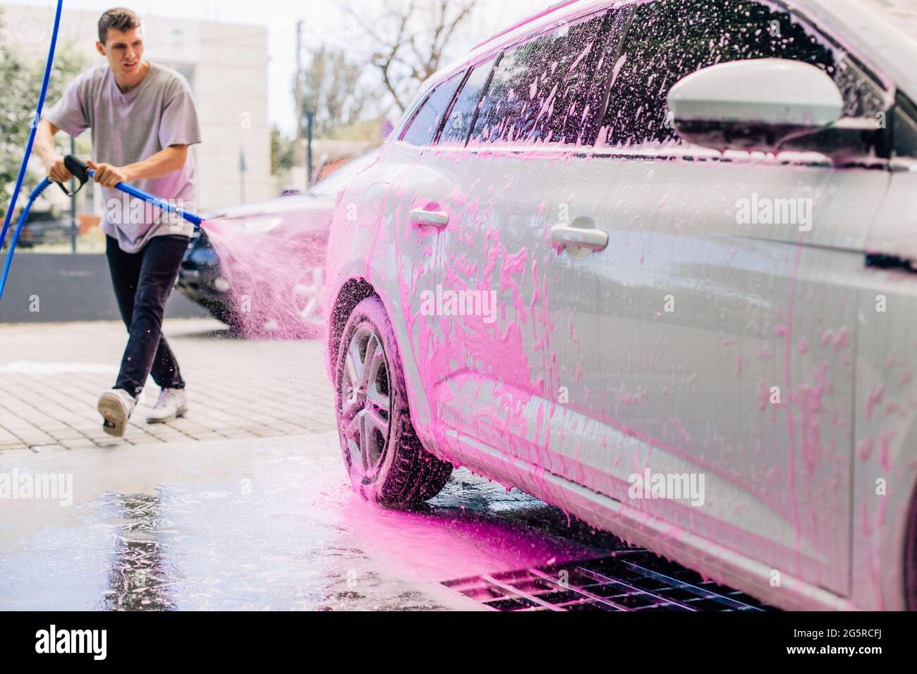 Young man washing his white car using pink foam to clean dirt, man washing off foam with high pressure water, Self-service concept, self-wash Stock Photo