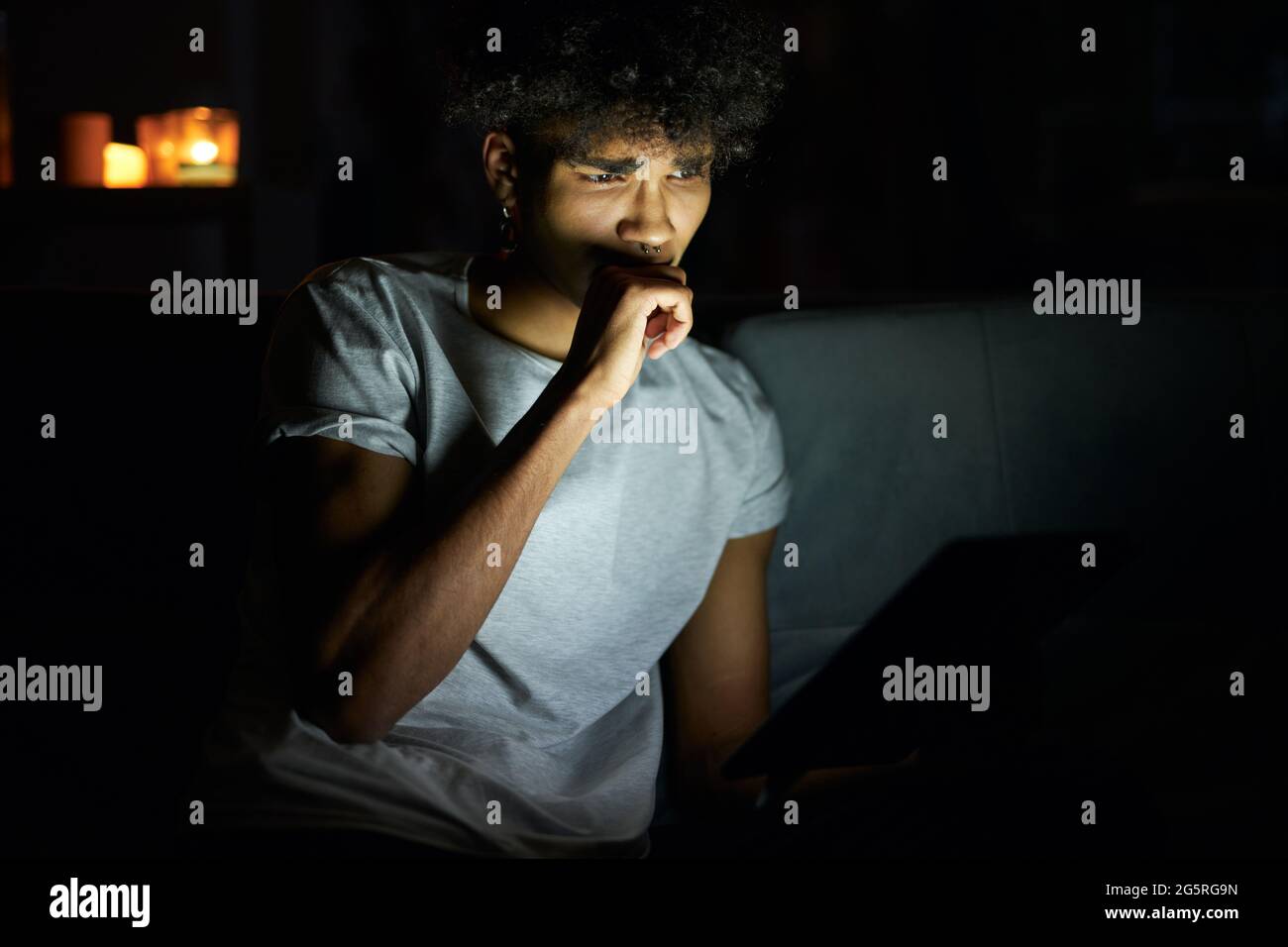 Portrait of young guy looking concentrated while reading e book using tablet pc at night, sitting in dark room at home Stock Photo