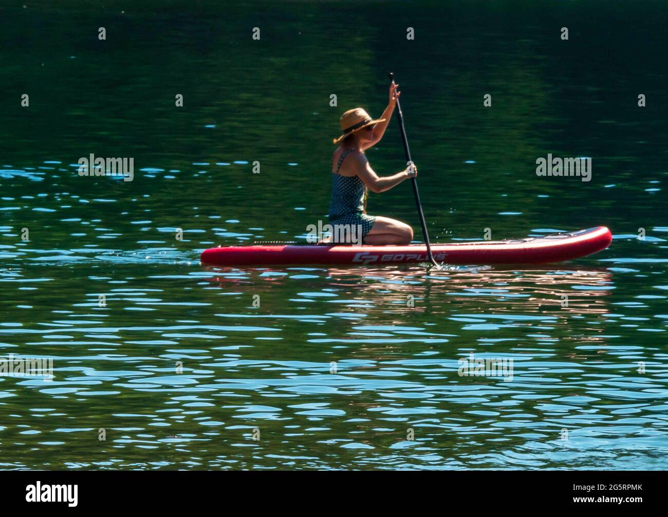 Young woman with hat, kneeling on a paddle board.  Green water, peaceful summer scene. Stock Photo