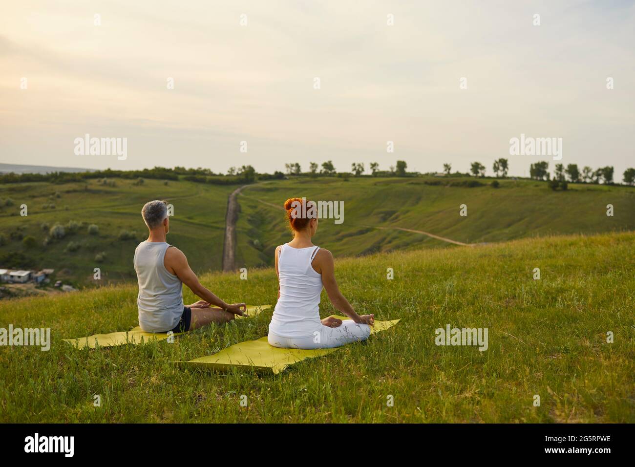 Couple sitting in Lotus pose and meditating on green hill on quiet summer evening in countryside Stock Photo